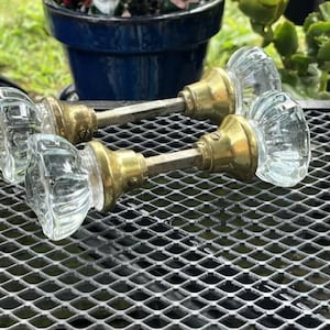May include: Two clear glass door knobs with faceted, star-shaped designs and brass-toned metal bases. Each knob is connected by a central metal spindle, showcasing a classic hardware style with transparent glass and metallic components.