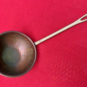 May include: A copper ladle with a long, slender handle and a hammered bowl. The bowl is round and has a dark, aged patina. The handle has a loop at the end for hanging. The ladle is set against a red background.