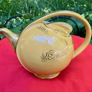 May include: A vintage yellow teapot with a gold handle, spout, and base. The teapot features gold floral and leaf designs. The teapot is sitting on a red surface, with green foliage in the background.