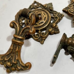 May include: Close-up of antique bronze drawer pulls with ornate floral and scrollwork designs. The hardware features a fleur-de-lis handle and a decorative circular backplate. The aged finish gives the pulls a vintage aesthetic.
