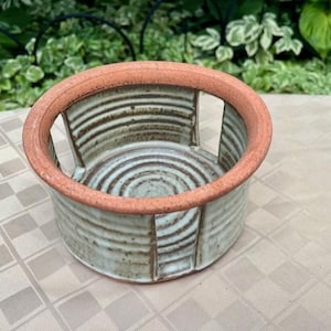May include: A round ceramic yarn bowl featuring a terracotta-colored rim and a light gray body with a ribbed, textured surface. The bowl includes a vertical cutout slot on the side for yarn to pass through, with a spiral pattern on the interior base.