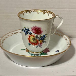 May include: White porcelain teacup and saucer set. The cup and saucer feature a floral design with red, yellow, and blue flowers. Both pieces have gold trim around the edges. The teacup is placed on top of the saucer.