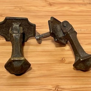 May include: Two antique bronze drawer pulls with a decorative design. Each pull has a rectangular backplate and a teardrop-shaped handle. A screw is attached to one of the pulls. The pulls are set against a wooden background.