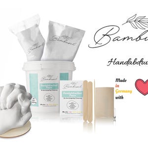 Könnte beinhalten: Ein weißes Handabdruck-Set mit einem Eimer Gips, zwei Beuteln Gips, zwei Holzstäbchen und einem herzförmigen Aufkleber mit der Aufschrift "Made in Germany with love".