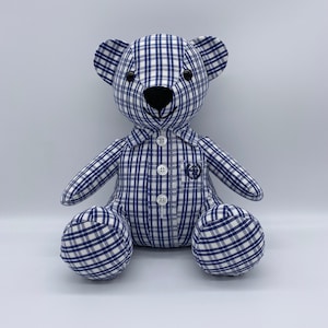 May include: A blue and white checked teddy bear with black eyes and a black nose. The bear is wearing a blue and white checked shirt with white buttons.