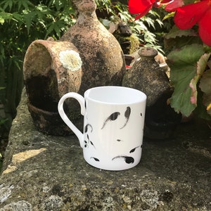May include: White ceramic mug with a curved handle, decorated with black abstract brushstrokes. The mug is set against a backdrop of weathered stone and greenery, suggesting an outdoor setting. The mug is a standard size.