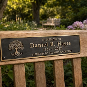 May include: A rectangular bronze memorial plaque with a dark finish and gold lettering, featuring an embossed tree icon. The text reads: IN MEMORY OF Daniel R. Hayes 1957 - 2022 A FRIEND TO ALL WHO KNEW HIM. The plaque is mounted on a wooden bench.