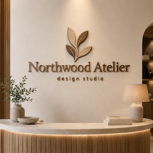 Può includere: Un'insegna a parete con il testo Northwood Atelier design studio in lettere marrone scuro e un'icona a forma di foglia in legno sopra. L'insegna è posizionata sopra un bancone reception curvo in legno con una lampada, libri e una pianta.