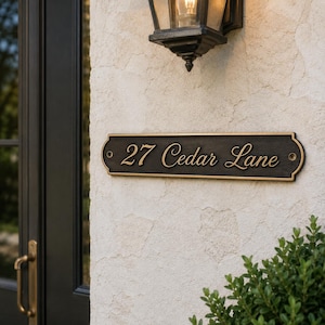 Può includere: Una targa indirizzo color bronzo con la scritta "27 Cedar Lane" in un elegante carattere. La targa è montata su un muro strutturato bianco sporco, vicino a una porta nera e una lampada decorativa per esterni. Un piccolo cespuglio verde si trova nell'angolo in basso a destra.