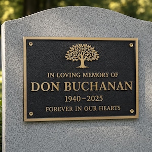 May include: A rectangular bronze memorial plaque mounted on a gray granite headstone. The plaque features a raised tree emblem, the text IN LOVING MEMORY OF DON BUCHANAN 1940-2025, and the phrase FOREVER IN OUR HEARTS, secured by four corner screws.
