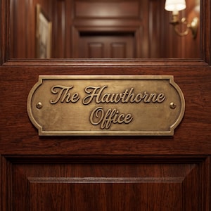 Könnte beinhalten: Ein Messing-Namensschild mit der Aufschrift "The Hawthorne Office" in eleganter Schrift ist an einer dunklen Holztür angebracht. Die rechteckige Platte hat abgerundete Kanten und ist mit kleinen Schrauben befestigt. Die Tür und die Holzverkleidung sind in einem satten, warmen Braunton gehalten.