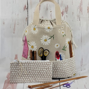 Op de afbeelding: Een beige ambachtelijke tas met madeliefjesprint en een bijpassend etui met stippen. De tas heeft een trekkoordsluiting en een crèmekleurige handgreep. Zakken bevatten een schaar, breinaalden en andere knutselspullen. Houten breinaalden en een paarse schaar zijn ook zichtbaar.