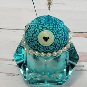 Pode incluir: Uma almofada de alfinetes de vidro turquesa com um topo redondo estampado. O topo é adornado com um colar de pérolas, botões e alfinetes decorativos. A base de vidro tem uma forma geométrica.