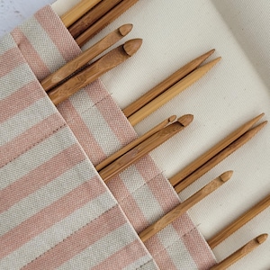 May include: A collection of wooden crochet hooks and knitting needles are stored in a striped pink and white fabric case. The hooks and needles are various sizes and are made of light brown wood. The case is open, revealing the tools.