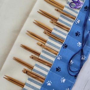 May include: A rolled-up craft organizer with wooden knitting needles and crochet hooks. The organizer has a blue and white striped section and a blue paw print patterned section. A black pen and purple scissors are also visible.