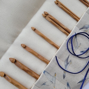 May include: A cream-colored crochet hook organizer with several wooden crochet hooks. The organizer has a flap with a blue bird pattern. A pair of purple scissors and a blue cord are also visible.