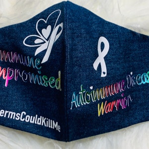 May include: A blue denim face mask with a white butterfly and ribbon design. The text "Immune Compromised" and "#YourGermsCouldKillMe" are printed in rainbow colors on the left side of the mask. The text "Autoimmune Disease Warrior" is printed in rainbow colors on the right side of the mask.