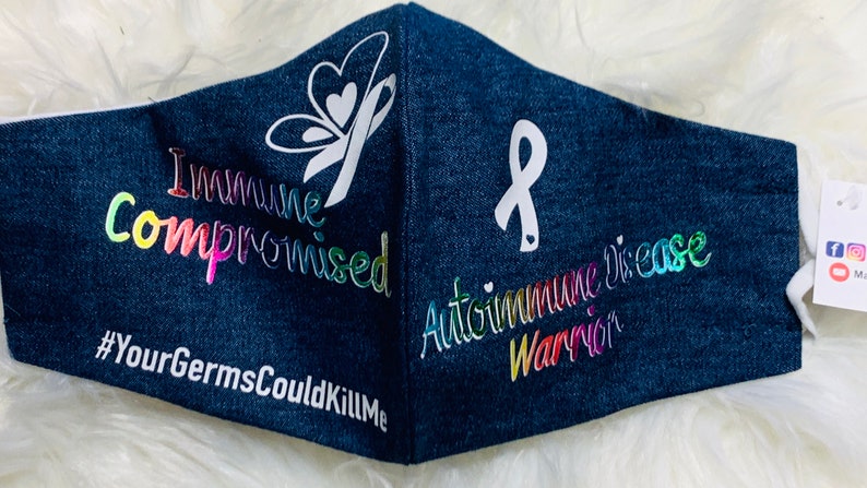 May include: A blue denim face mask with a white butterfly and ribbon design. The text "Immune Compromised" and "#YourGermsCouldKillMe" are printed in rainbow colors on the left side of the mask. The text "Autoimmune Disease Warrior" is printed in rainbow colors on the right side of the mask.