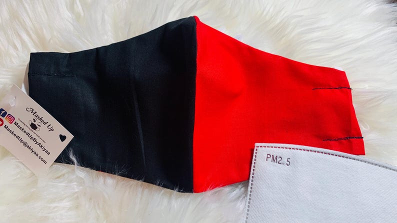 May include: Black and red face mask with a white filter insert labeled "PM2.5". The mask is made of two fabrics, one black and one red, sewn together. The mask has a small tag attached with the text "Masked Up" and a website address.