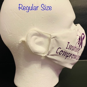 May include: White face mask with purple text that reads "Immune Compromised" and a purple ribbon symbol. The mask is on a white mannequin head. The text "Regular Size" is written in blue above the mask.
