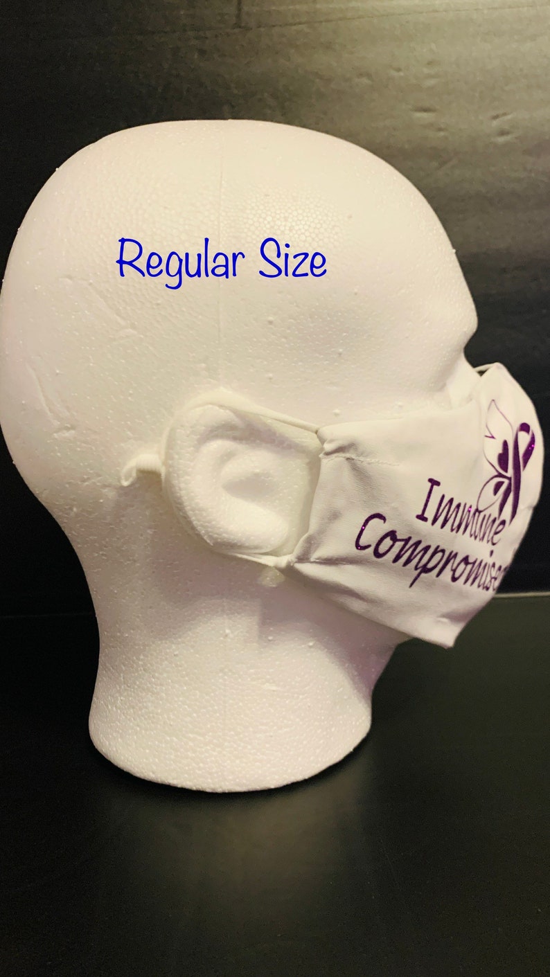May include: White face mask with purple text that reads "Immune Compromised" and a purple ribbon symbol. The mask is on a white mannequin head. The text "Regular Size" is written in blue above the mask.