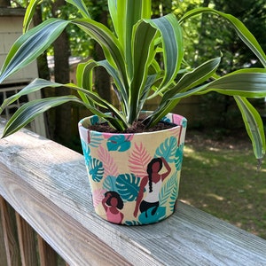 Puede incluir: Una planta verde en una maceta de tela con un estampado tropical con mujeres. El estampado es amarillo, rosa y azul con hojas verdes.