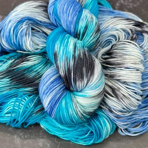 May include: A skein of hand-dyed yarn in shades of blue, teal, and black. The yarn is tightly wound in a skein, with visible variations in color and texture. Ideal for knitting, crocheting, or other fiber arts projects.