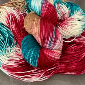 May include: A skein of hand-dyed yarn featuring a variegated color palette of deep teal, vibrant magenta, soft cream, and warm tan. The fibers are twisted into a central bundle with loose, flowing strands extending outward in all directions.