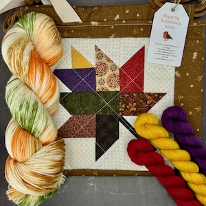 May include: A fabric project bag featuring a quilted maple leaf design in autumn colors, accompanied by a large variegated yarn skein in green, orange, and white, plus three smaller solid yarn skeins in red, yellow, and purple.