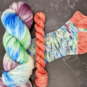 May include: A close-up of colorful yarn skeins and a knitted sock. The yarn is in shades of blue, green, purple, and white. A smaller skein of coral-colored yarn is also present, along with a sock with a similar color palette.