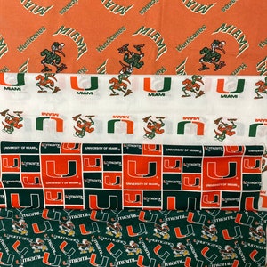 May include: Three different fabric swatches featuring the University of Miami Hurricanes logo. The first swatch is orange with a white logo and a cartoon mascot. The second swatch is white with a green and orange logo. The third swatch is green with an orange logo and a cartoon mascot.