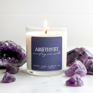 May include: A lit candle in a clear glass jar with a navy blue label that reads "AMETHYST coconut soy wax candle." The candle is surrounded by several amethyst crystals and geodes. The candle is 10 oz.