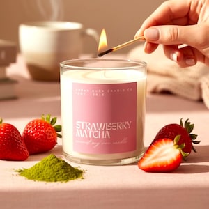 May include: A lit candle in a clear glass jar with a pink label that reads "Strawberry Matcha". Fresh strawberries and matcha powder are arranged around the candle. A match is being used to light the candle. A cup and books are in the background.