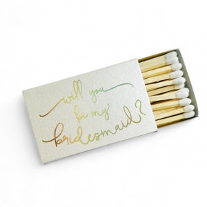 May include: A white matchbox with gold foil lettering that reads "Will you be my bridesmaid?" The matchbox is filled with wooden matches with white tips.