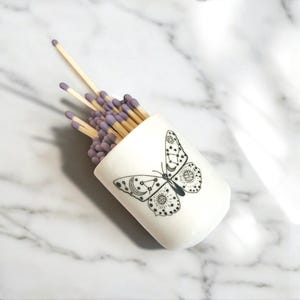 May include: A white ceramic match striker with a black and white butterfly design. The butterfly has celestial details, including moons, stars, and suns. The striker is filled with matches with purple tips.