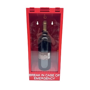 Może przedstawiać: Czerwone, prostokątne pudełko z przezroczystym panelem przednim, przeznaczone do przechowywania butelki wina. Tekst "BREAK IN CASE OF EMERGENCY" jest wydrukowany na dole. Wewnątrz znajduje się ciemna butelka wina ze złotą szyjką.