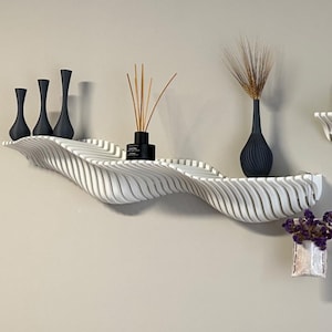 Puede incluir: Dos estanterías de pared blancas en forma de onda con jarrones negros, plantas secas y una pequeña planta en maceta. Las estanterías están montadas en una pared gris claro, creando una estética moderna y minimalista. Las estanterías miden unos 100 cm de largo.
