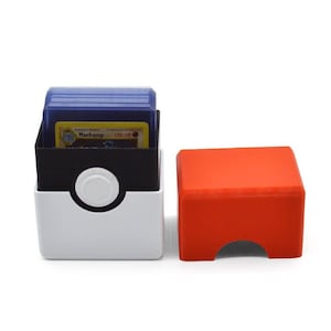 May include: A white and black Pokemon card storage box with a red lid. The box has a Pokeball design on the front and is open to reveal a stack of blue Pokemon cards. The card on top of the stack is a Machhamp card with 100 HP.