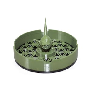 May include: A green ceramic ashtray with a Yoda figure in the center. The ashtray has a flower of life design around the edge.