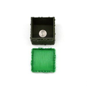 Puede incluir: Una caja de pl&aacute;stico verde y marr&oacute;n con tapa. La caja tiene forma de castillo y tiene una superficie texturizada. La tapa est&aacute; abierta y hay una moneda de veinticinco centavos dentro de la caja.