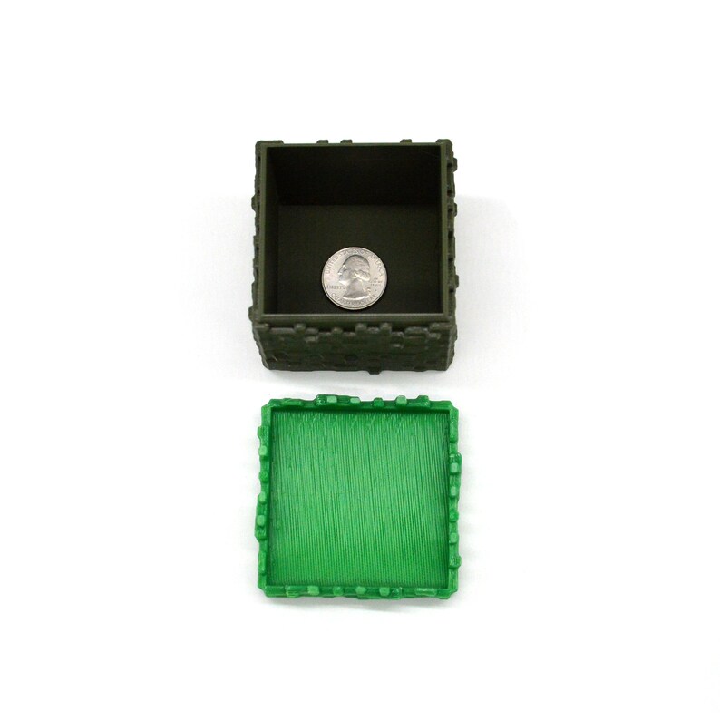 Puede incluir: Una caja de pl&aacute;stico verde y marr&oacute;n con tapa. La caja tiene forma de castillo y tiene una superficie texturizada. La tapa est&aacute; abierta y hay una moneda de veinticinco centavos dentro de la caja.