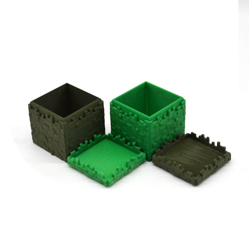 Puede incluir: Dos peque&ntilde;as cajas cuadradas con tapas. Una caja es verde y la otra es marr&oacute;n. Ambas cajas tienen una superficie texturizada que se asemeja a bloques de piedra.