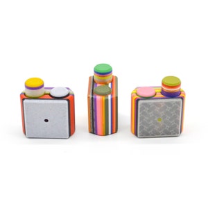 May include: Three colourful, striped, cube-shaped objects with a small, round, coloured button on top of each. The cubes are made of layered, colourful stripes. The buttons are yellow, green, and pink.