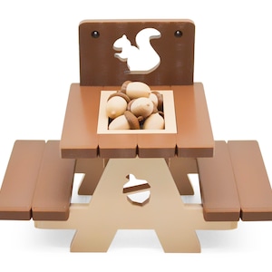 May include: A brown wooden children's picnic table with a squirrel cutout on the back. The table has a built-in centre compartment filled with acorn-shaped toys.