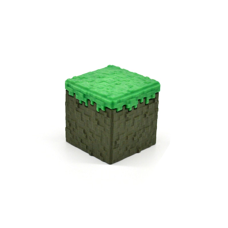 Puede incluir: Un bloque de juguete de pl&aacute;stico verde y marr&oacute;n con forma de bloque de hierba de Minecraft.