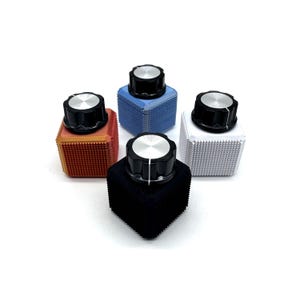 Pode incluir: Uma coleção de cinco botões coloridos com tampas prateadas. Os botões são montados em bases quadradas texturizadas em tons de laranja, azul, branco e preto. Os botões estão dispostos sobre um fundo branco.