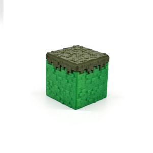 Puede incluir: Un bloque de juguete de pl&aacute;stico verde y marr&oacute;n con forma de bloque de hierba de Minecraft.