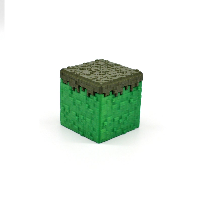 Puede incluir: Un bloque de juguete de pl&aacute;stico verde y marr&oacute;n con forma de bloque de hierba de Minecraft.