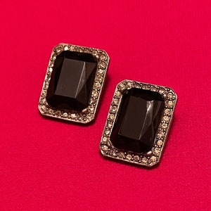 Puede incluir: Un par de pendientes de piedra negra con marco de metal plateado. Las piedras preciosas son rectangulares y tienen un diseño facetado.