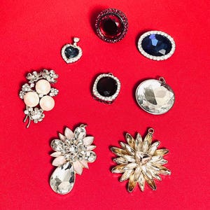 May include: A collection of sparkling jewelry pieces, including brooches, pendants, and decorative stones, arranged on a red surface. The jewelry features various shapes, colors, and designs, including a heart-shaped pendant, a floral brooch, and several large, faceted stones.